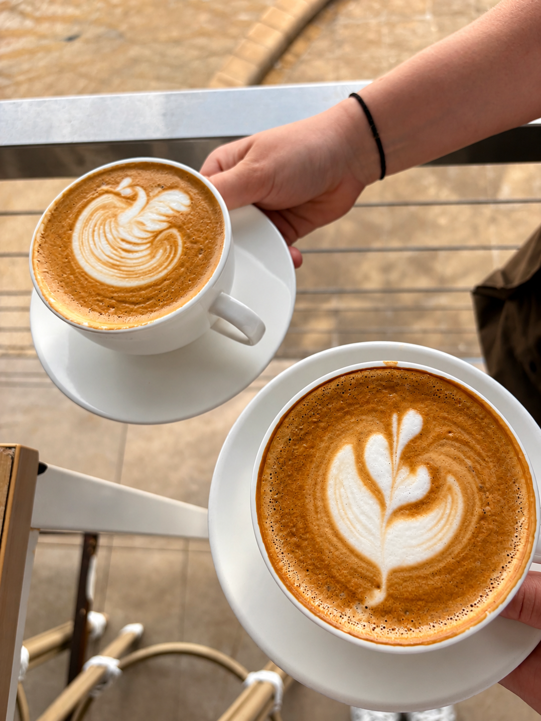 Two lattes with beautiful latte art held by guests at an outdoor table